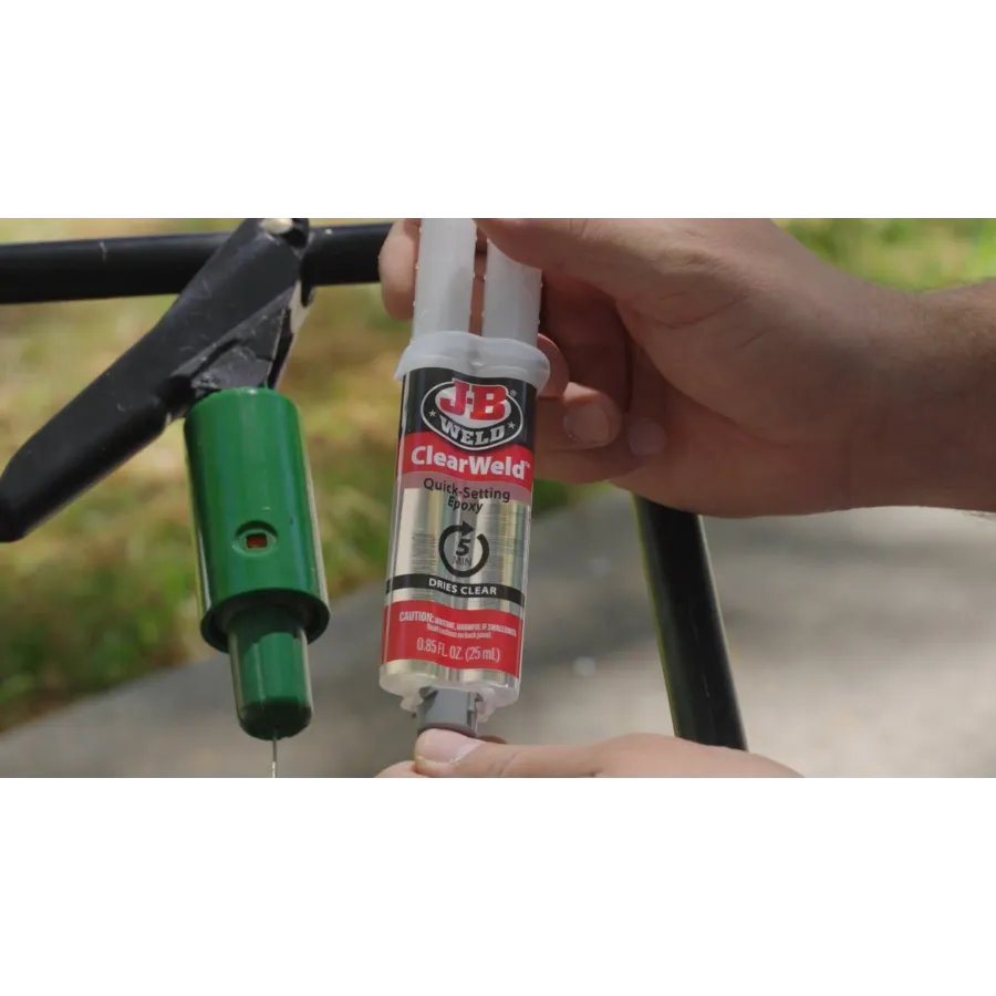 Hands dispensing JB Weld ClearWeld quick-setting epoxy for repair of a green metal object outdoors.