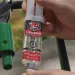 Hands dispensing JB Weld ClearWeld quick-setting epoxy for repair of a green metal object outdoors.