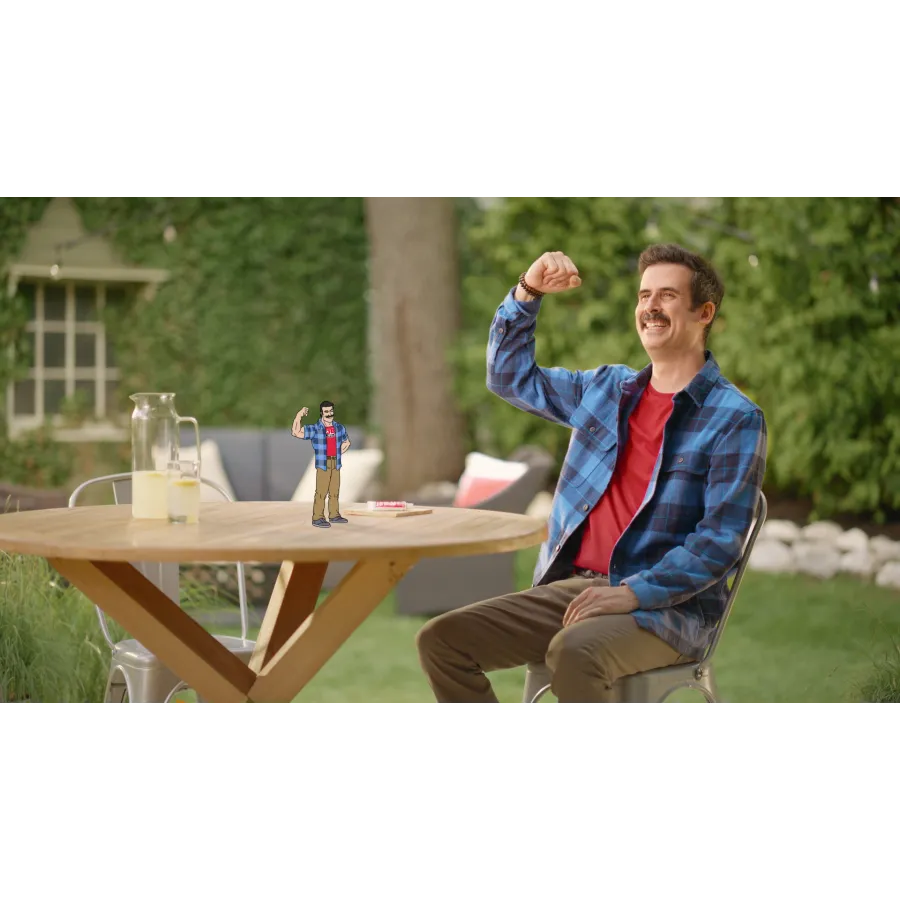 A man in a flannel shirt and mustache waves joyfully at a tiny animated character on a garden table.