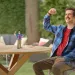 A man in a flannel shirt and mustache waves joyfully at a tiny animated character on a garden table.